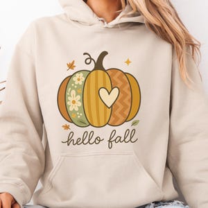May include: Beige hoodie featuring a pumpkin design with floral and heart patterns, and the words "hello fall". The hoodie has a front pocket and a hood. The design is in autumnal colours.