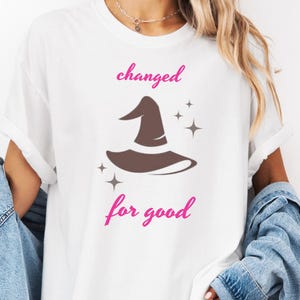 May include: White t-shirt featuring the text "changed for good" in pink script, with a brown witch hat graphic. The shirt has a relaxed fit and rolled-up sleeves, suitable for casual wear.