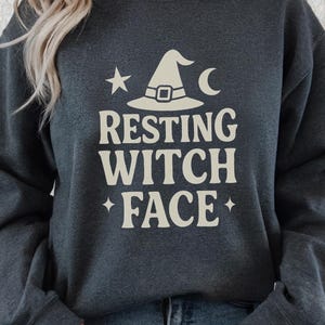 May include: Dark gray sweatshirt with the text "RESTING WITCH FACE" in a cream color. Above the text is a witch hat, star, and crescent moon graphic. The sweatshirt is a casual, long-sleeved garment.