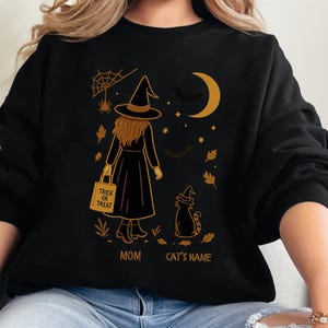 May include: Black Halloween sweatshirt featuring a witch design. The graphic includes a witch with a trick-or-treat bag, a cat in a witch hat, a moon, bats, and the text "MOM" and "CAT'S NAME".