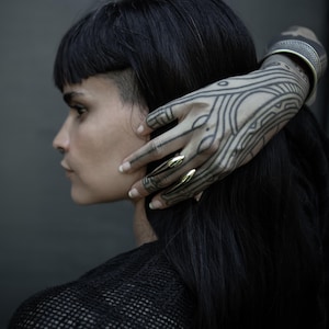 May include: A side profile of a person with black hair and gold-colored nail covers. The person's hand, adorned with intricate black tattoos, rests on their head. A silver bracelet is visible on the wrist.