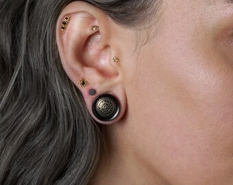 May include: A gold ear cartilage stud with four black stones, a gold eye-shaped stud, a small three-bead gold stud, a gold flower-shaped stud with a black center, and a large black ear plug featuring a circular gold filigree pattern on the front.