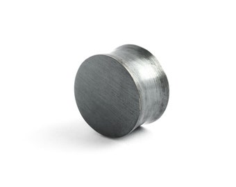 May include: A cylindrical ear plug with a dark, brushed metallic finish. The circular front face is flat with a visible grain texture, while the sides of the plug curve inward toward the center, creating a smooth, concave profile in dark gray.