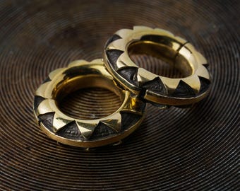 May include: A pair of circular ear gauges featuring a polished gold-tone outer rim with a serrated, sunburst-like edge. The inner section of each gauge is finished in a dark, oxidized metal with a textured, etched pattern visible on the surface.