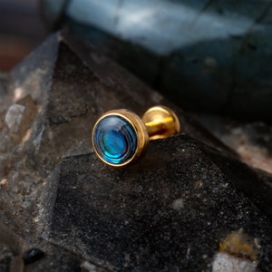 Peut inclure: Un bijou de corps en or avec une incrustation de nacre d'ormeau bleue. Le bijou est un clou circulaire avec une tige.