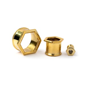 May include: Three gold-colored, hexagonal-shaped body jewelry pieces. The largest piece is a tunnel, the middle piece is a plug, and the smallest is a stud. All pieces have a shiny, metallic finish and are set against a white background.
