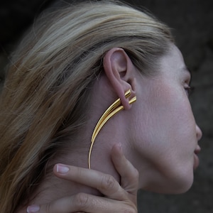 May include: A pair of gold ear cuffs with a curved, pointed design. The cuffs are worn in the upper ear, with one cuff extending down the side of the neck.