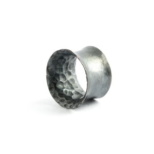 Puede incluir: Un expansor de oreja de metal gris oscuro con una abertura ancha y acampanada. La superficie interior presenta una textura martillada distintiva con pequeñas hendiduras redondeadas, mientras que la banda exterior tiene un acabado mate cepillado.