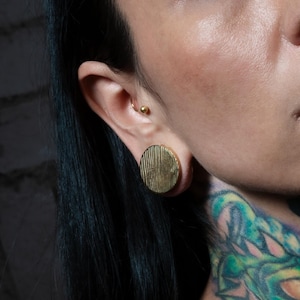 Puede incluir: Un expansor de oreja circular de tono dorado con una textura de rayas verticales en su superficie frontal plana. La joya está insertada en un lóbulo estirado y se observa un pequeño pendiente de bola dorado en el cartílago superior.
