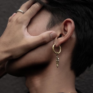 Könnte beinhalten: Ein goldener Creolenohrring mit einem baumelnden, strukturierten Pfeil-Anhänger.