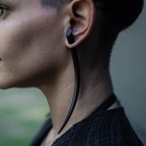 May include: A black ear gauge, or ear stretcher, is worn in the ear. The gauge has a cylindrical top and a curved, tapered design. The jewellery is inserted into the earlobe and extends downwards. The gauge is made of a dark material.