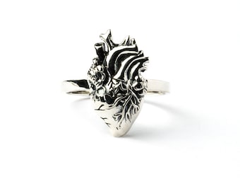 May include: A silver-toned ring featuring a detailed, three-dimensional anatomical heart sculpture as the centerpiece. The metal surface has oxidized recessed areas to highlight the intricate veins, arteries, and chambers of the heart design.
