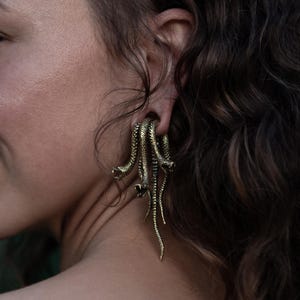 May include: Gold-coloured earrings with a snake design. The earrings feature multiple snake heads and dangling bodies, creating a unique accessory. The jewellery is worn on the ear, adding a bold statement to any look.