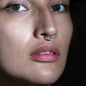 May include: Close-up of a person's face with a silver nose ring. The person has dark brown eyes and full lips.