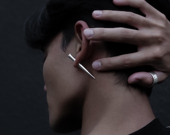 Könnte beinhalten: Ein silberfarbener Ohrring in Form eines spitzen Kegels, der durch das Ohrläppchen gesteckt ist. Der Ohrring hat eine glatte, polierte metallische Oberfläche, die das Licht reflektiert und eine scharfe, spitz zulaufende Silhouette erzeugt.