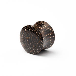 May include: A dark brown wooden ear plug with a speckled pattern on the front and a curved shape. The plug has a natural wood grain with lighter streaks. The product is set against a white background.