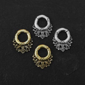 May include: Four ornate ear gauges in gold and silver tones. Each gauge features a circular design with a hinged opening and a decorative filigree base. The gauges are displayed on a dark, textured surface.