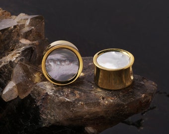 May include: A pair of gold-toned ear plugs with a cylindrical shape. One plug features a circular inlay with a dark, swirling, marbled pattern, while the other plug displays a smooth, iridescent white mother-of-pearl circular inlay.