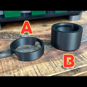 May include: Two black, 3D-printed plastic components sit on a wooden surface. One is a ring with a curved cutout, and the other is a cylindrical cup. The letter "A" is next to the ring, and the letter "B" is next to the cup.