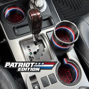May include: Car interior with cup holders and a gear shift. The cup holders have red, white, and blue trim with a tire track design. The text "PATRIOT EDITION" is displayed in a patriotic color scheme.
