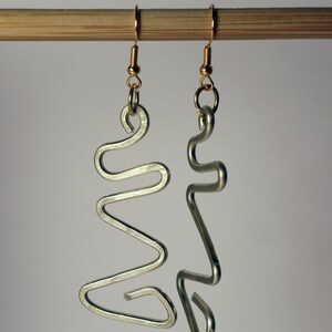 Könnte beinhalten: Ein Paar silberfarbene, abstrakte Drahtohrringe. Jeder Ohrring hat ein einzigartiges, fließendes Design mit goldfarbenen Ohrhaken. Die Ohrringe werden vor einem neutralen Hintergrund präsentiert, was ihre moderne, minimalistische Ästhetik hervorhebt.