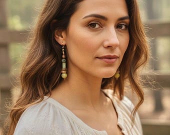 May include: A woman wearing long, dangling earrings with green and brown beads. The earrings are suspended from gold-colored hooks. She is wearing a cream-colored blouse with embroidered details. Her long, wavy brown hair frames her face.