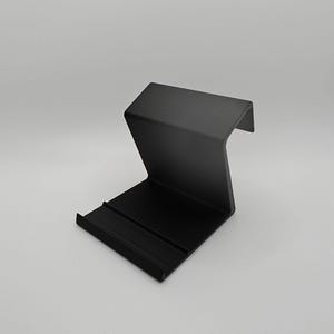 May include: A black plastic phone stand with a raised back and a flat front. The stand is designed to hold a phone upright.