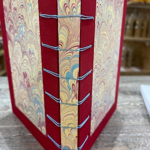 Könnte beinhalten: Ein handgefertigtes Notizbuch mit rotem Einband und marmorierten Papierseiten. Die Seiten sind mit hellblauem Faden gebunden. Das marmorierte Papier weist Wirbel aus Gelb, Rot und Blau auf. Die Ränder des Journals sind ebenfalls rot.