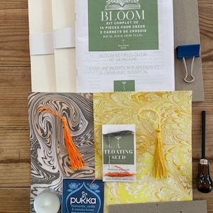 Può includere: Un kit artigianale con una varietà di materiali, tra cui carta, un libretto con la scritta "BLOOM" e una bustina di tè. Il kit include una clip blu, uno strumento per intagliare e carta decorativa con nappe arancioni e gialle.