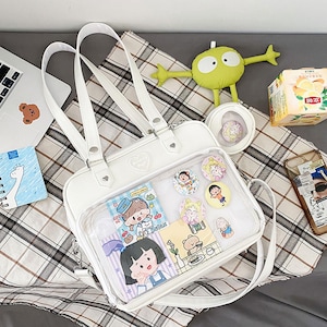 Puede incluir: Bolso escolar blanco de pl&aacute;stico transparente y piel sint&eacute;tica con un bolsillo de malla y un bolsillo frontal transparente. El bolsillo frontal tiene una variedad de pegatinas y pines con personajes de dibujos animados. El bolso tiene un asa superior y una correa para el hombro.