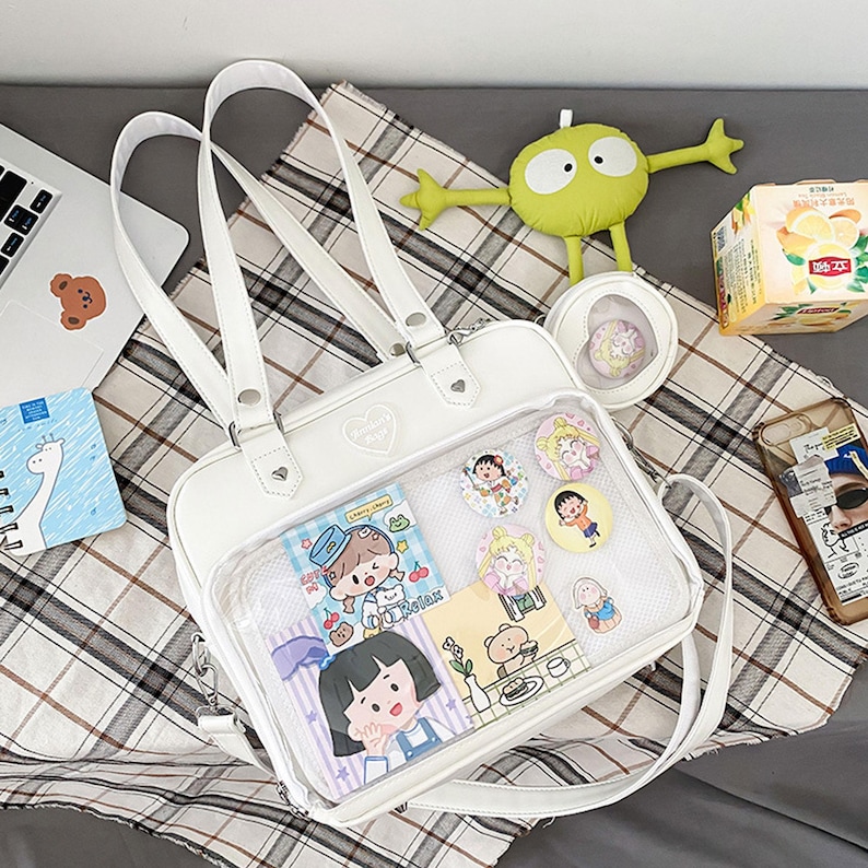 Puede incluir: Bolso escolar blanco de pl&aacute;stico transparente y piel sint&eacute;tica con un bolsillo de malla y un bolsillo frontal transparente. El bolsillo frontal tiene una variedad de pegatinas y pines con personajes de dibujos animados. El bolso tiene un asa superior y una correa para el hombro.