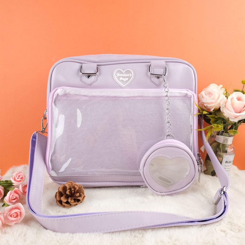 Puede incluir: Un bolso de hombro de color lavanda con un bolsillo frontal transparente de pl&aacute;stico y un dije en forma de coraz&oacute;n. El bolso tiene una correa de cadena plateada y un logotipo en forma de coraz&oacute;n plateado que dice "Brooklyn's Bagz".