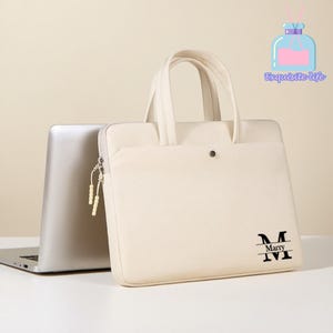 Könnte beinhalten: Cremefarbene Laptoptasche mit Griff und Reißverschluss. Auf der Tasche steht der Text "Marry" in einer schwarzen, stilisierten Schriftart. Ein silberner Laptop ist teilweise hinter der Tasche zu sehen. Die Tasche hat einen Knopfverschluss.