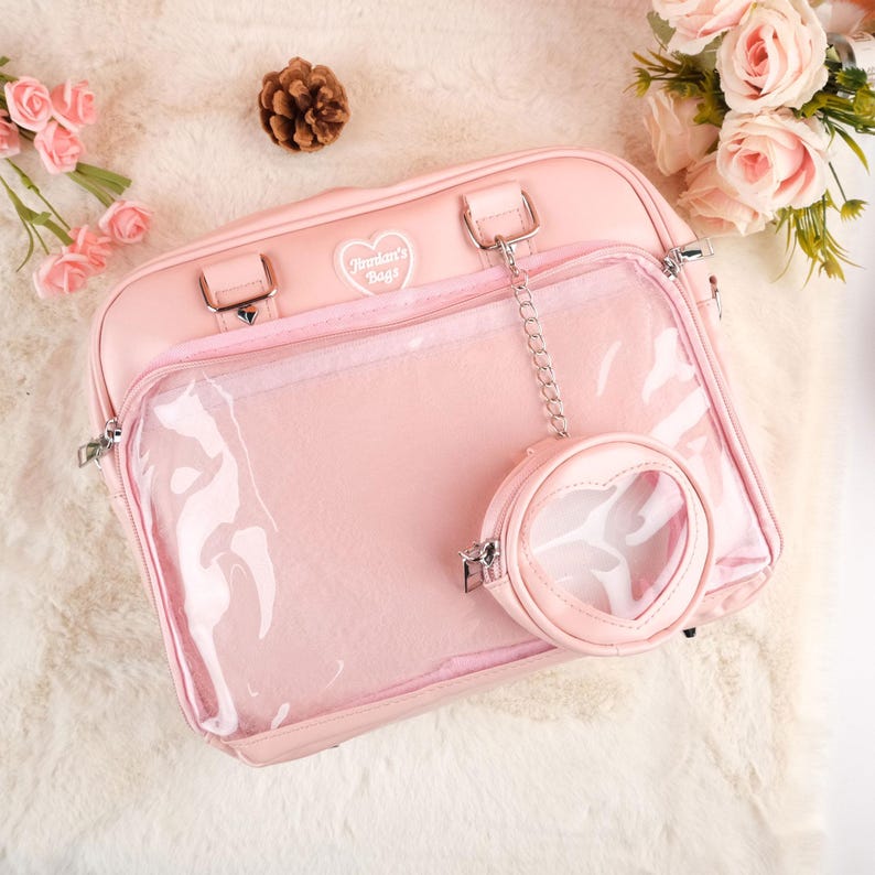 Puede incluir: Un bolso rosa de pl&aacute;stico transparente y piel sint&eacute;tica con un dije en forma de coraz&oacute;n. El bolso tiene un panel frontal de pl&aacute;stico transparente y un panel trasero de piel sint&eacute;tica rosa. El bolso tiene un logotipo bordado en blanco que dice "Blossom's Bags".
