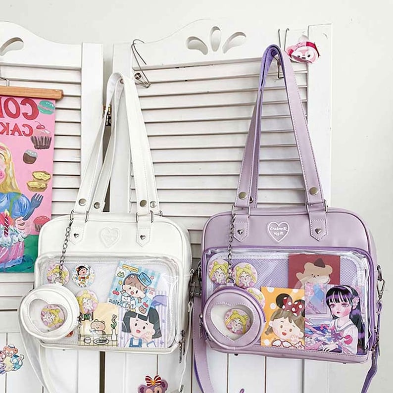 Puede incluir: Dos bolsas de mano transparentes de pl&aacute;stico con ribetes de imitaci&oacute;n de cuero blanco y morado. Las bolsas tienen un dije en forma de coraz&oacute;n y un detalle de cadena. La bolsa blanca tiene un bolsillo transparente con calcoman&iacute;as de dibujos animados y la bolsa morada tiene un bolsillo transparente con calcoman&iacute;as de dibujos animados y un peque&ntilde;o espejo redondo.
