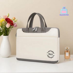 May include: A cream laptop bag with grey trim and handles, featuring a top handle and a zip closure. The bag has a decorative wreath design with the name "Marry" printed on the front. A vase of flowers and a perfume bottle are next to the bag.