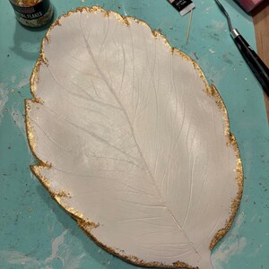 Pode incluir: Uma bandeja branca em forma de folha com texturas de veios detalhadas e bordas douradas com acabamento em flocos de ouro metálico. A superfície é lisa e fosca, contrastando com a aplicação texturizada e cintilante de folha de ouro.