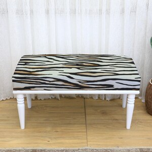 May include: A small bench with a zebra print seat cushion and white legs. The cushion features a pattern of black, brown, and cream stripes. The bench is a decorative furniture piece.