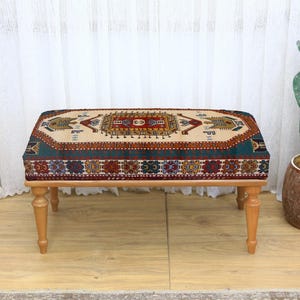 May include: A rectangular ottoman with a patterned top and wooden legs. The top features a beige background with a central design in red, brown, and blue. The legs are light brown, and the overall style is traditional.