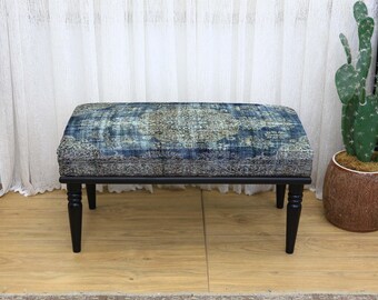 May include: A rectangular ottoman with a patterned blue and gray upholstered top and black wooden legs. The ottoman is placed on a light wood floor, with a cactus in a brown pot to the right.
