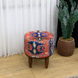 Op de afbeelding: Een ronde, rood, blauw en wit gepatroneerde poef met vier houten poten. De poef staat op een houten vloer.