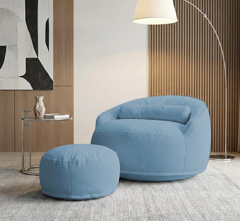May include: A light blue corduroy beanbag chair with a matching ottoman. The chair has a rounded back and a cylindrical pillow. The ottoman is circular. The furniture is in a modern living room setting.