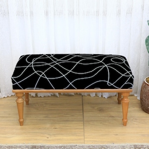 May include: A small wooden bench with a black seat featuring a white, abstract, squiggly line design. The bench has four light brown, turned legs. A cactus in a brown pot is to the right.