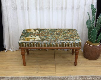 Può includere: Un pouf rettangolare in legno con un piano in tessuto fantasia nei toni del verde, giallo e marrone. Ha quattro gambe tornite e poggia su un pavimento in legno. Un cactus in un vaso marrone è a destra. Dimensioni: circa 70 cm di lunghezza.