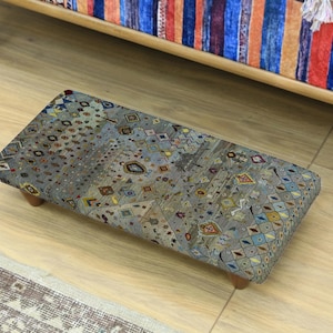 May include: A rectangular footstool with a patterned fabric top in shades of grey, blue, and yellow. The footstool has four short, wooden legs. The fabric features a repeating geometric design.
