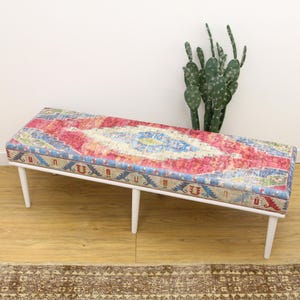 Peut inclure: Un banc blanc avec un coussin à motifs colorés. Le coussin présente un motif géométrique rouge, bleu et blanc. Le banc est posé sur un sol en bois.