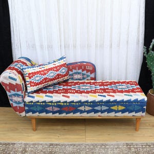 May include: A small, patterned sofa with a matching pillow. The sofa features a wooden frame and legs. The upholstery is a mix of red, blue, and white geometric patterns. A cactus in a brown pot sits to the right.