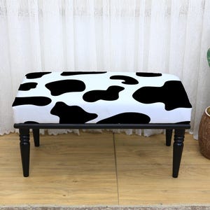 May include: A rectangular bench with a black frame and legs. The seat is upholstered in a white fabric with large black cow spots. The bench is approximately 36 inches long and 18 inches wide.