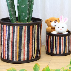 Puede incluir: Dos cestas de almacenamiento de tela a rayas, una más grande que la otra. La cesta más grande está llena de una planta de cactus. La cesta más pequeña contiene un oso de peluche marrón y un conejo de peluche blanco. Un león de peluche marrón y blanco está en primer plano.