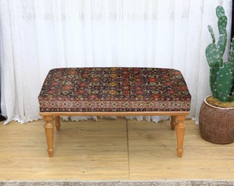 May include: A rectangular ottoman with a patterned cushion in shades of red, brown, and gold. The wooden legs are light brown. The ottoman is placed on a light wood floor, with a cactus in a brown pot in the background.